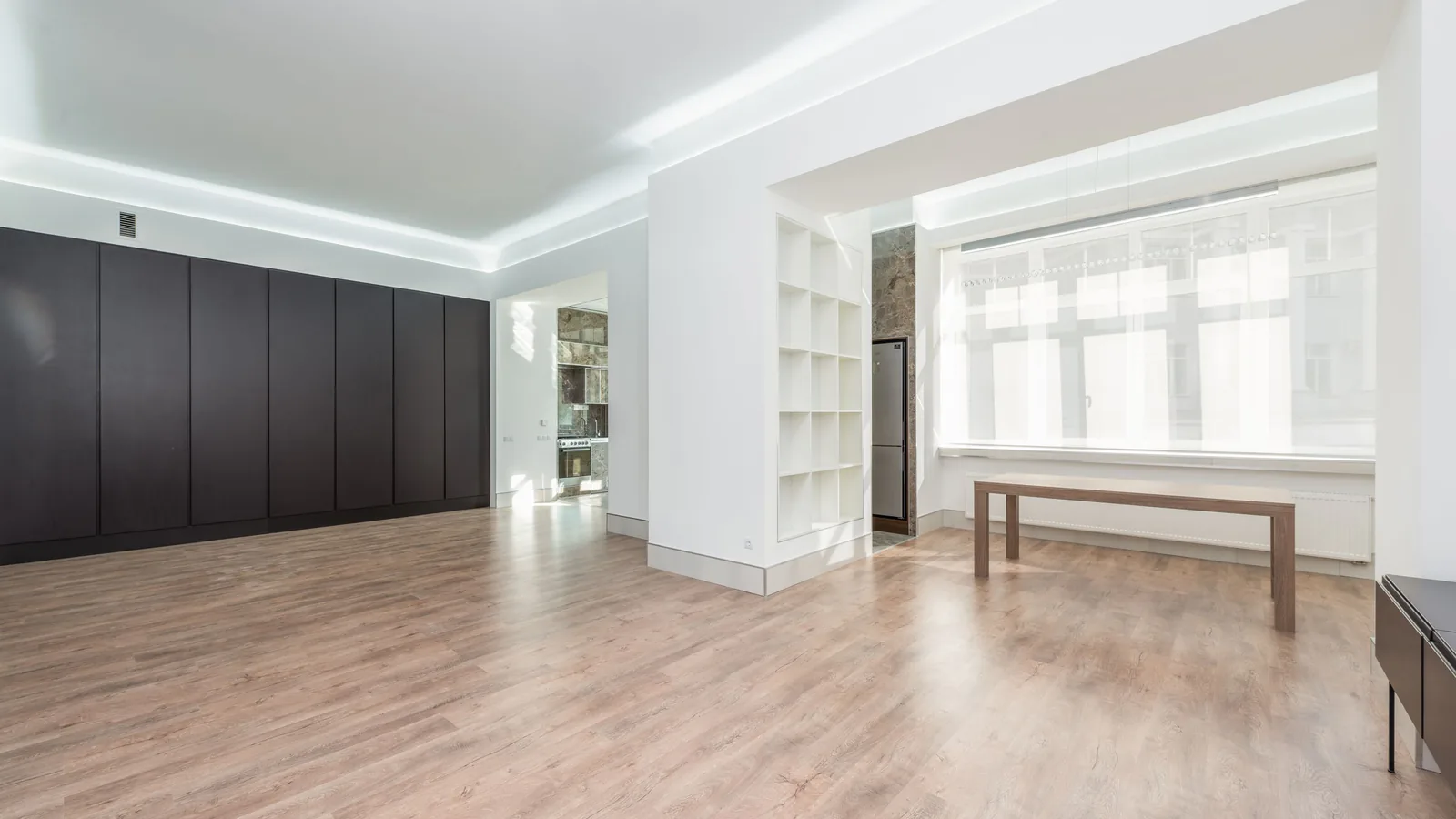 An empty room with pale wood flooring, built in shelving and a long table under a run of windows.